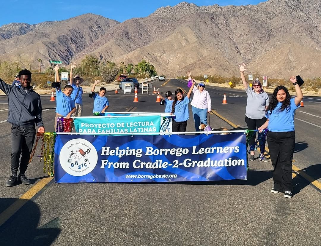 Students and volunteers carrying a BASIC banner down a closed road at a Borrego Springs parade, mountains behind them