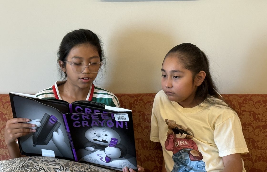 An older Learning Academy student reads a picture book aloud to a younger student