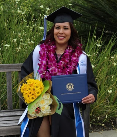A BASIC scholar in cap and gown holding her CSU San Marcos diploma and a bouquet