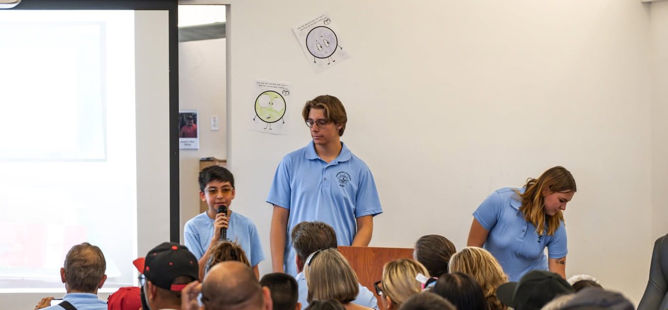 A BASIC Learning Academy student speaking into a microphone to a seated audience, with two program staff beside him