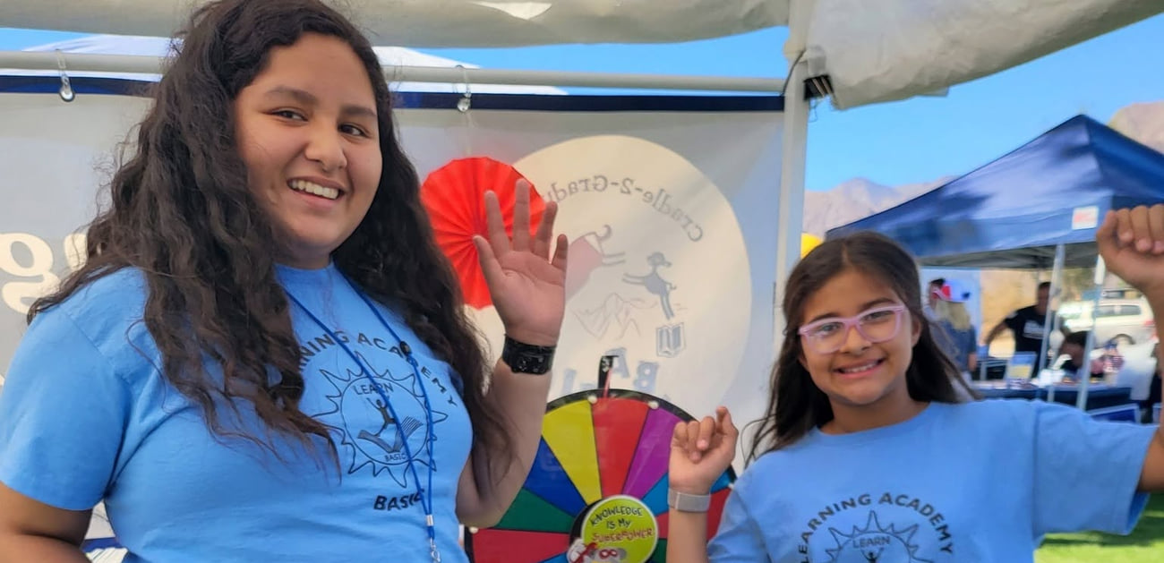 Two BASIC Learning Academy students staffing a prize-wheel booth at an outdoor community event, with the Borrego Springs mountains behind them